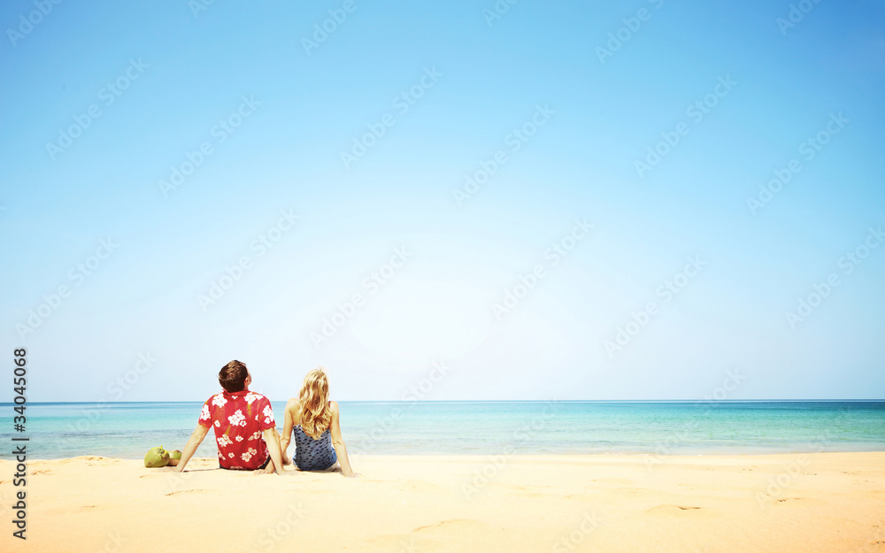 Young couple sitting on the sand and looking to a blue tropical sky