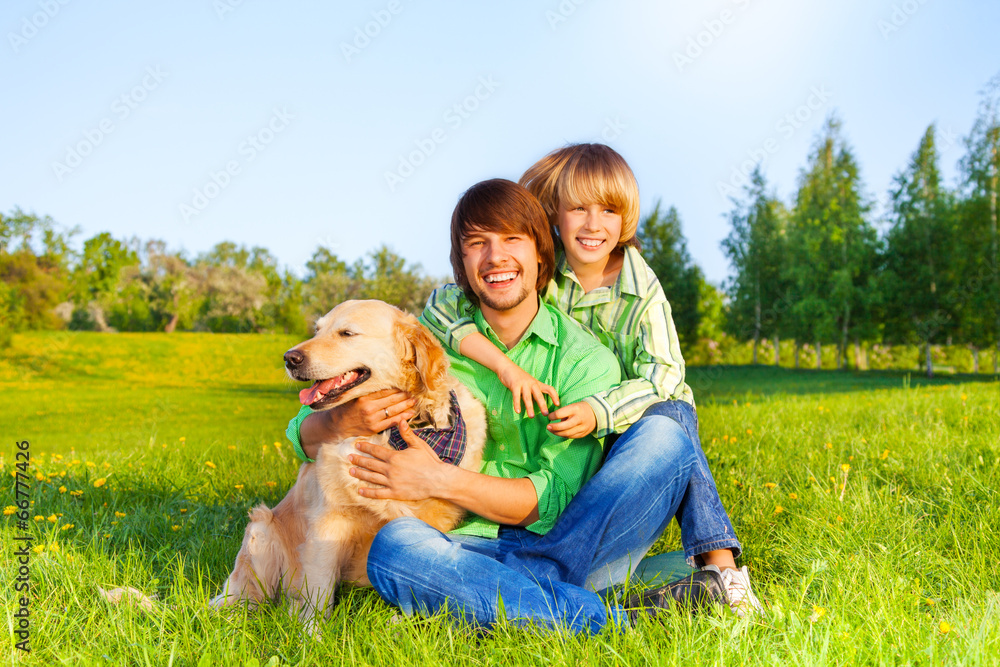 Smiling father, kid  and dog sit in park on grass