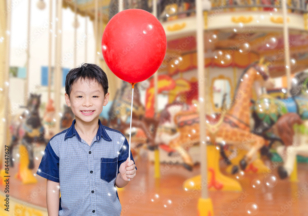 Asian boy holding red balloon in front of merry go round