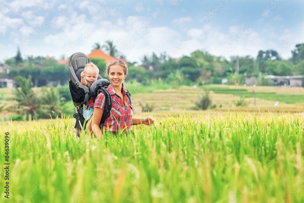 Cheerful mother with baby boy in carrying backpack walking on green rice terraces. Traveling with ch