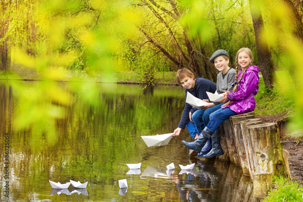 Smiling friends sit putting paper boats on pond