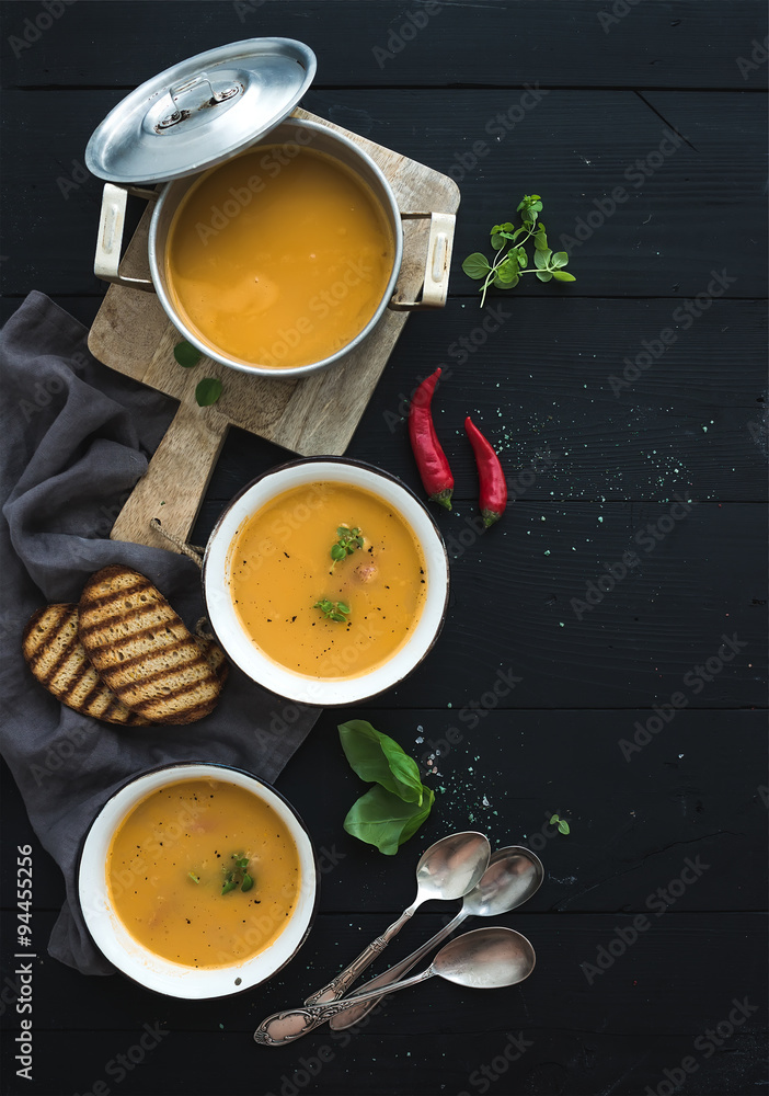 Red lentil soup with spices, herbs, bread in a rustic metal saucepan and bowls, over dark wood backd