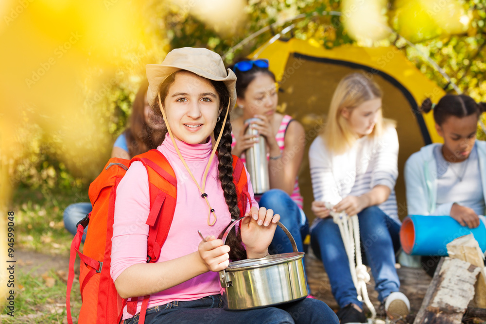 Smiling girl wears hat, holds metallic pot at camp