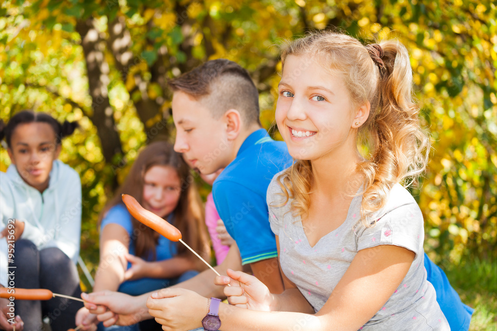 Girl and teens sit on campsite with sausages