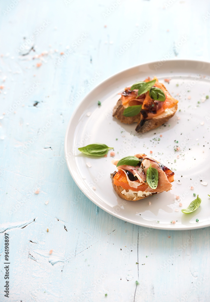 Bruschettas with Prosciutto，烤甜瓜，软奶酪和罗勒放在白色陶瓷盘子上，在灯光下