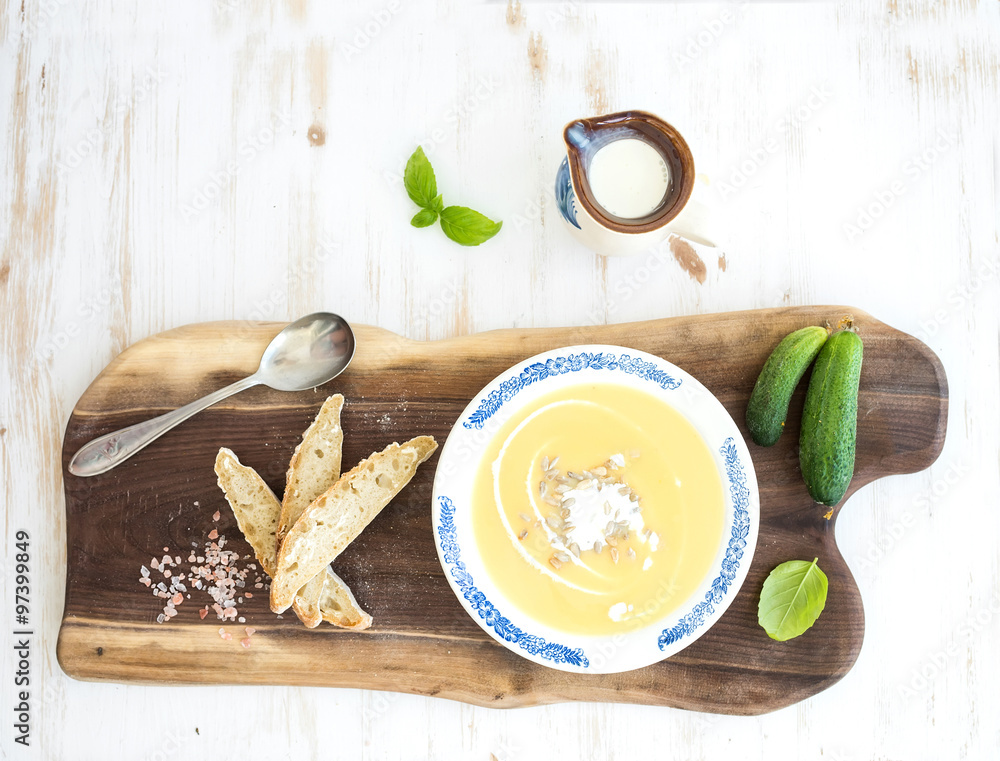 Pumpkin soup with cream, fresh basil, cucumbers and bread in vintage ceramic plate on wooden board o