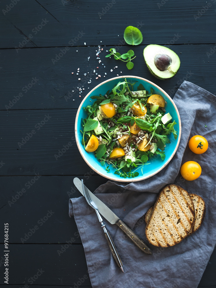 Bowl of green salad with avocado, arugula, cherry tomatoes and sunflower seeds, grilled bred slices,