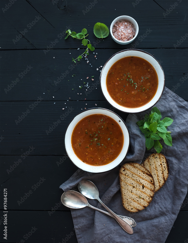 Roasted tomato soup with fresh basil, spices and bread in rustic metal bowls over black background