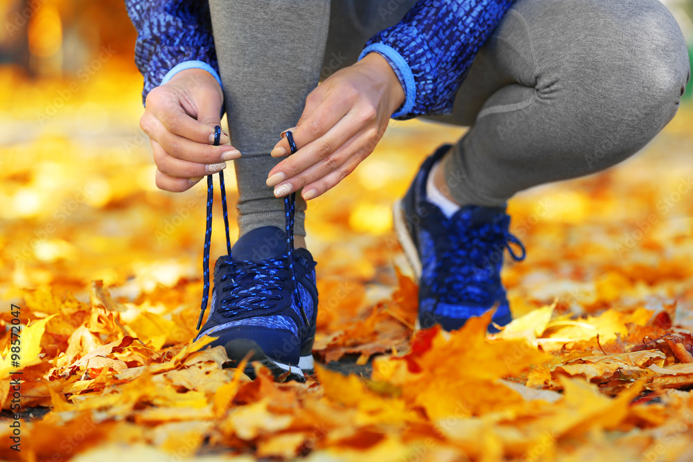 Young woman tying laces on her sport shoes before training in autumn forest.