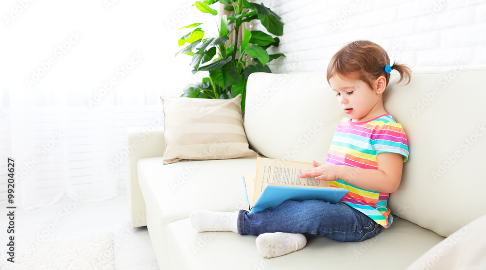 cute baby girl reading book at home sitting on sofa