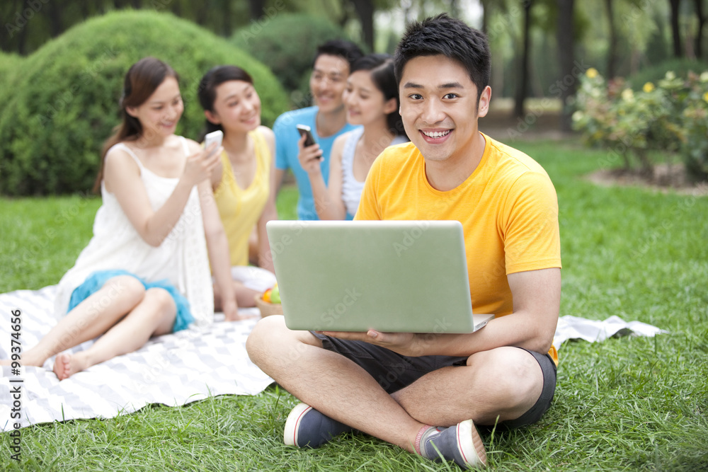 Young man using computer on meadow with friends in background