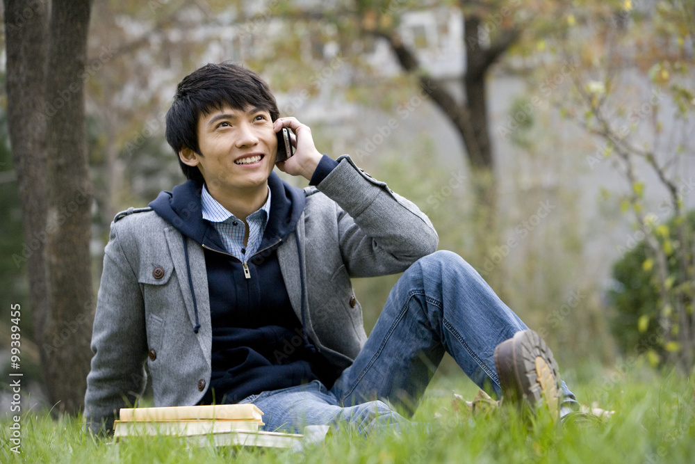 A young man sitting on a grassy area talking on the phone