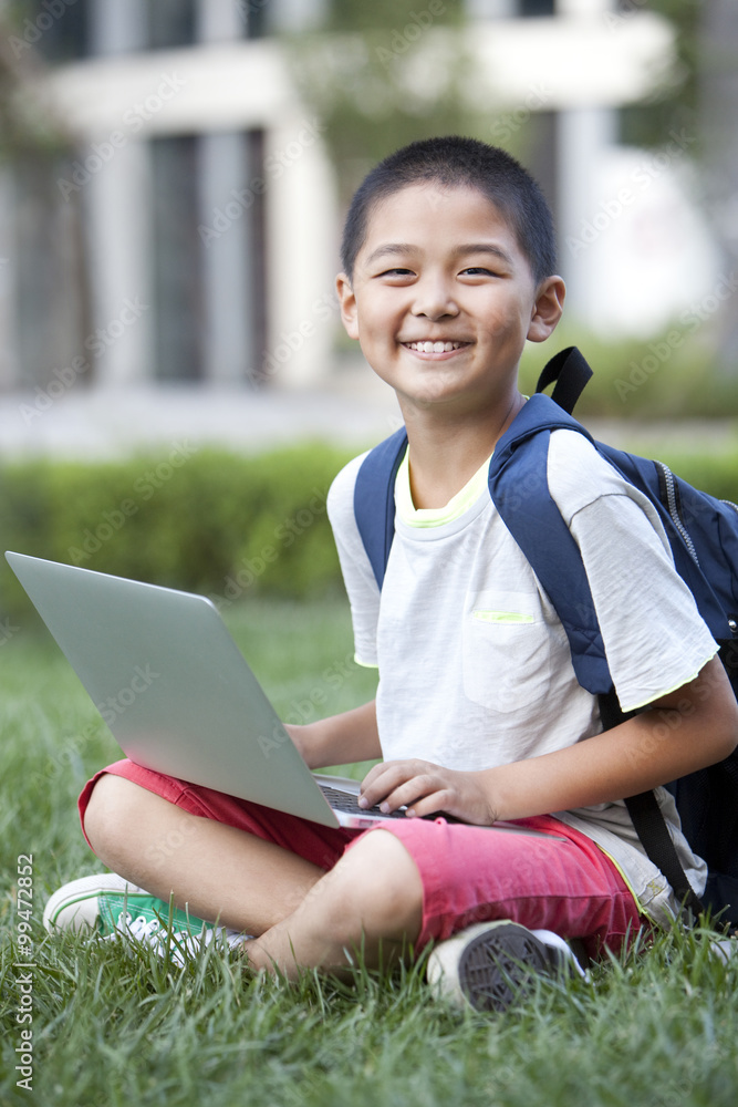 Smiling schoolboy sitting on the lawn with laptop