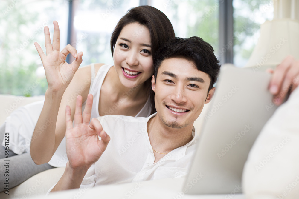 Cheerful young couple doing OK sign with digital tablet in living room