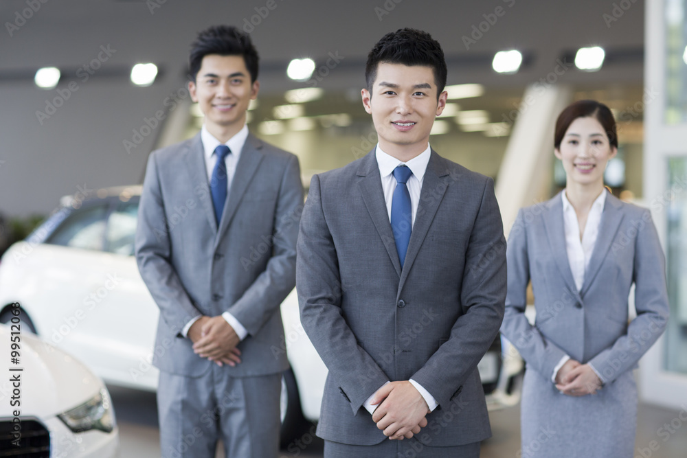 Confident salespeople standing with new cars in showroom