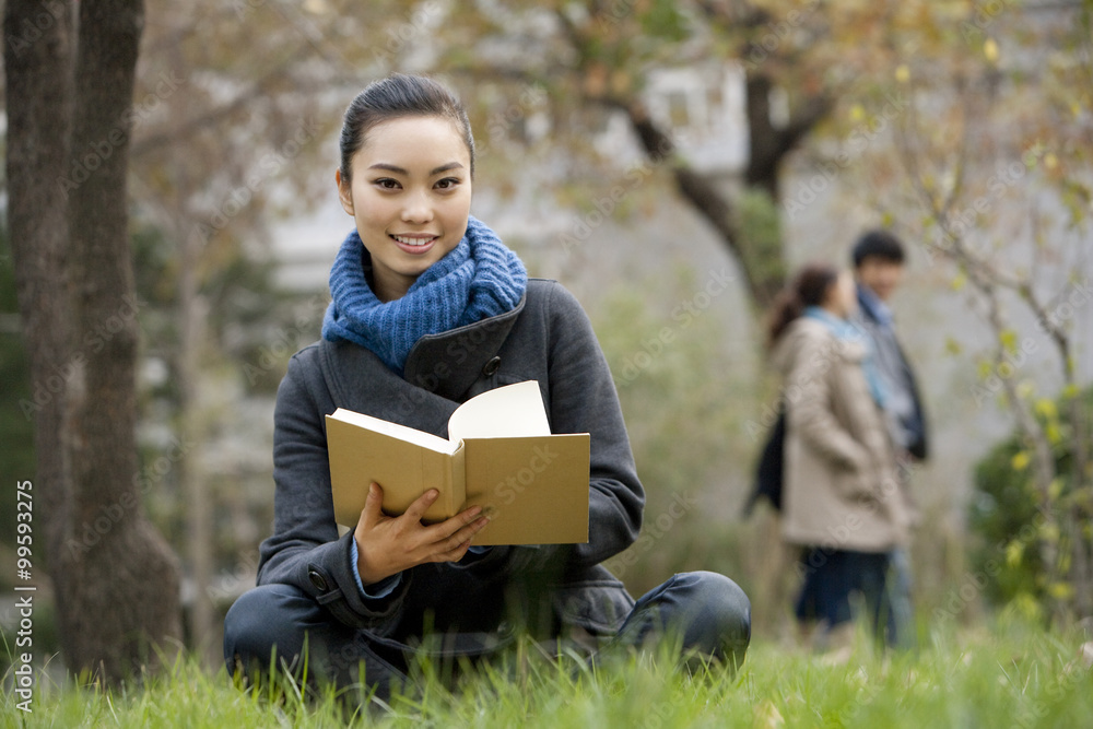 A young woman sitting on a grassy area reading a book, people in the background