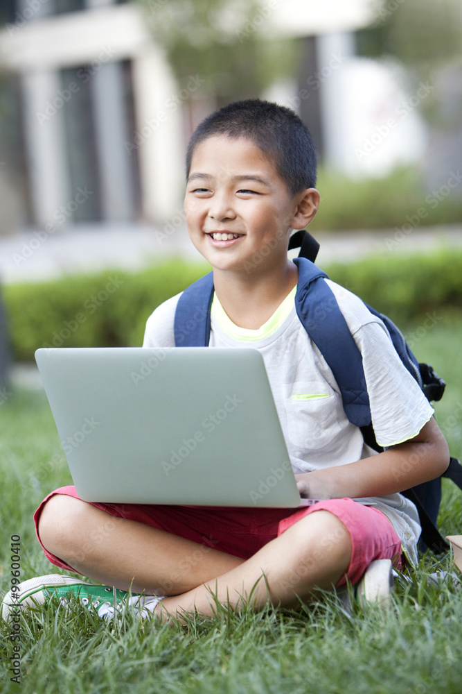 Smiling schoolboy sitting on the lawn with laptop