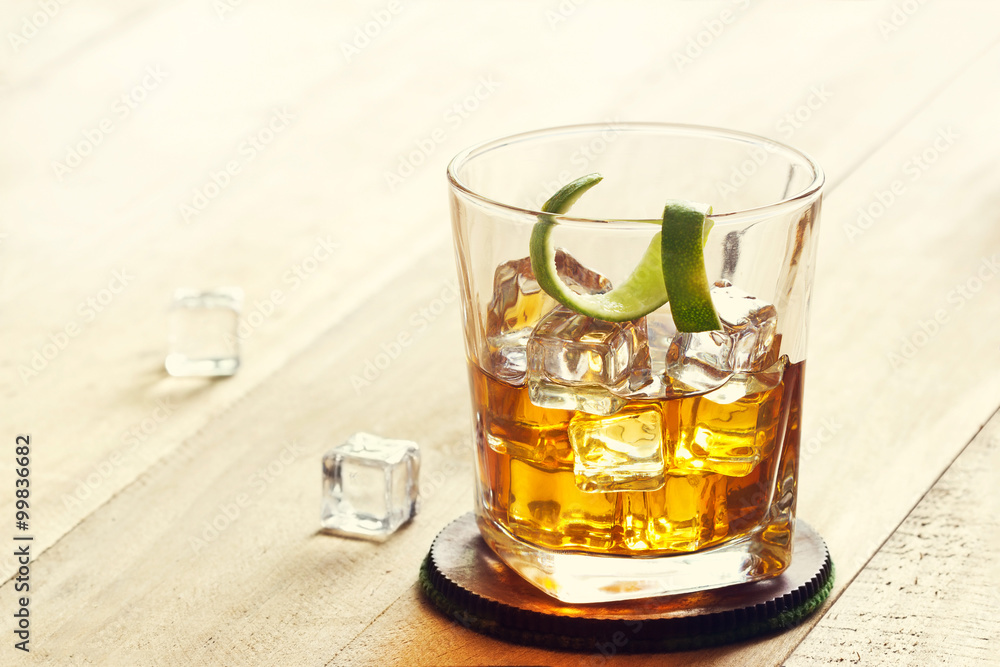 Glass of whiskey with ice and lime peel on wooden background, warm color tone