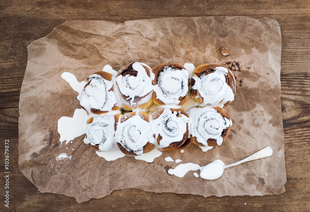 Cinnamon rolls with cream cheese icing on piece of oily craft paper over rustic wooden background