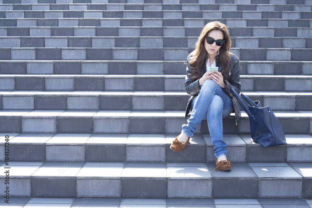 Young woman with sunglasses is looking at the smartphone sitting on the stairs