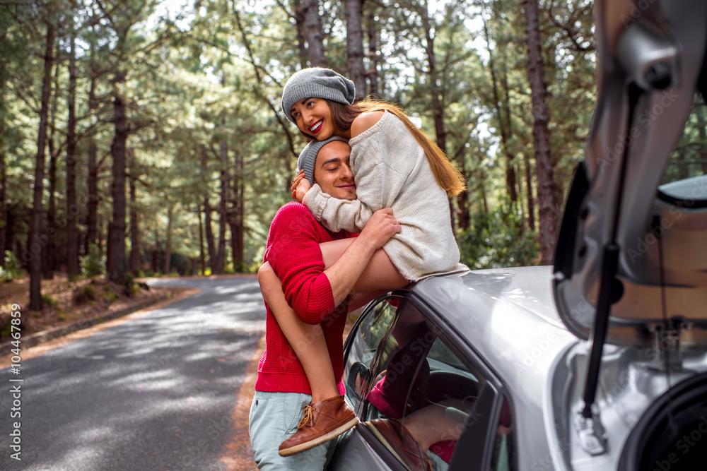 Young and lovely couple in sweates and hats having fun hugging together near the car on the roadside