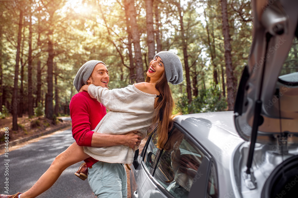 Young and lovely couple in sweates and hats having fun hugging together near the car on the roadside