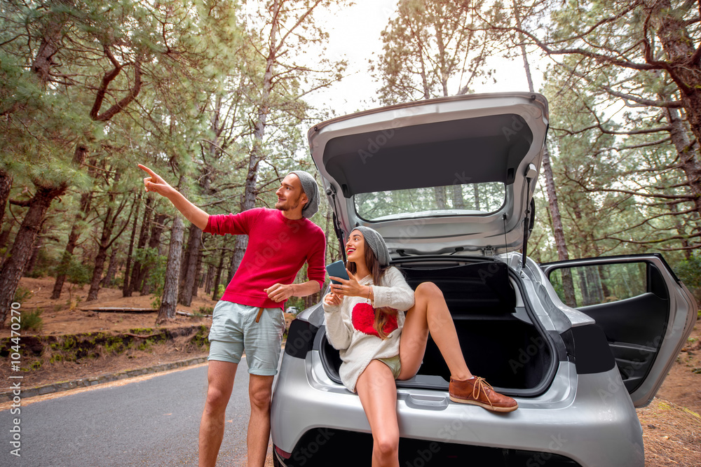 Pretty couple in sweaters and hats sitting in the car trunk on the forest roadside. Young family tra