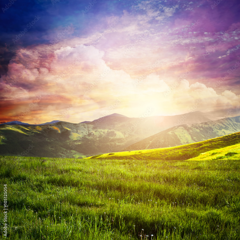 Fairtytale landscape with green grass, mountains, sunset fantastic sky.