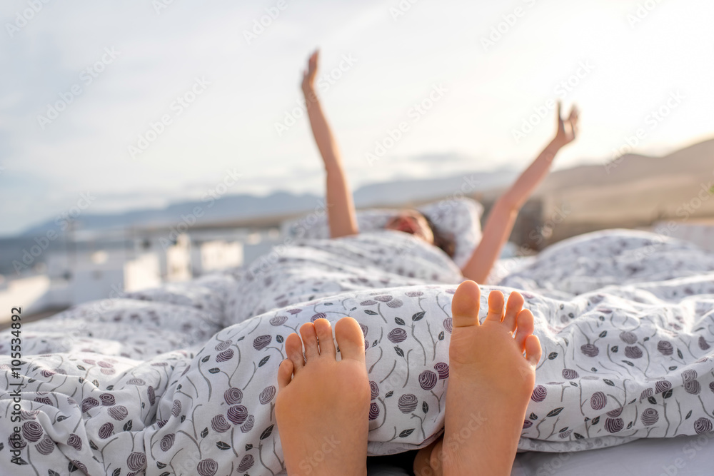 Young and cute woman waking up lying on the bed on the roof top with white houses on the background.