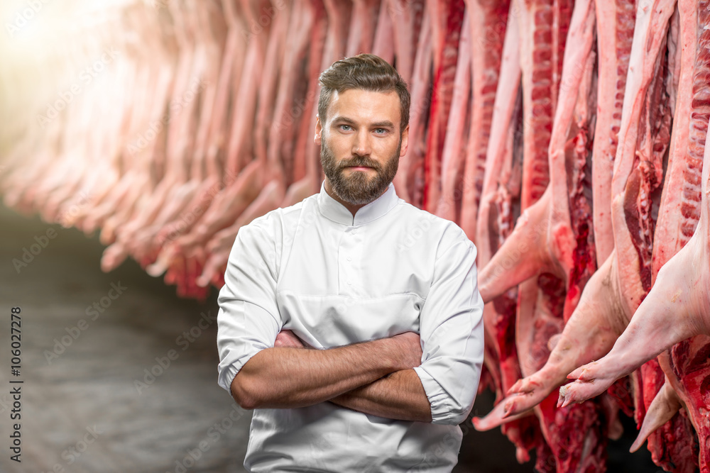 Portrait of a handsome butcher in white uniform at the meat manufacturing with pork carcasses on the