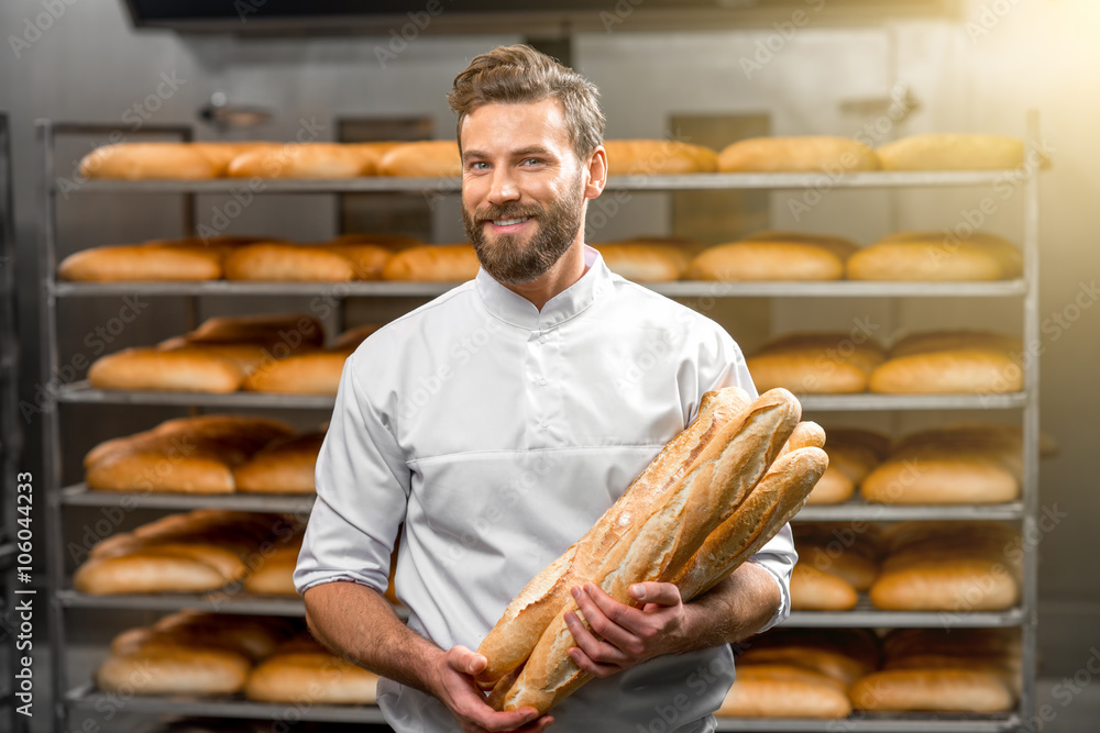 Handsome baker in uniform holding baguettes with bread shelves on the background at the manufacturin