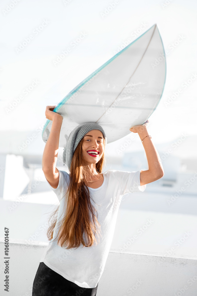 Portrait of a young smiling woman dressed casual carrying surfboard on the white city background. Ac