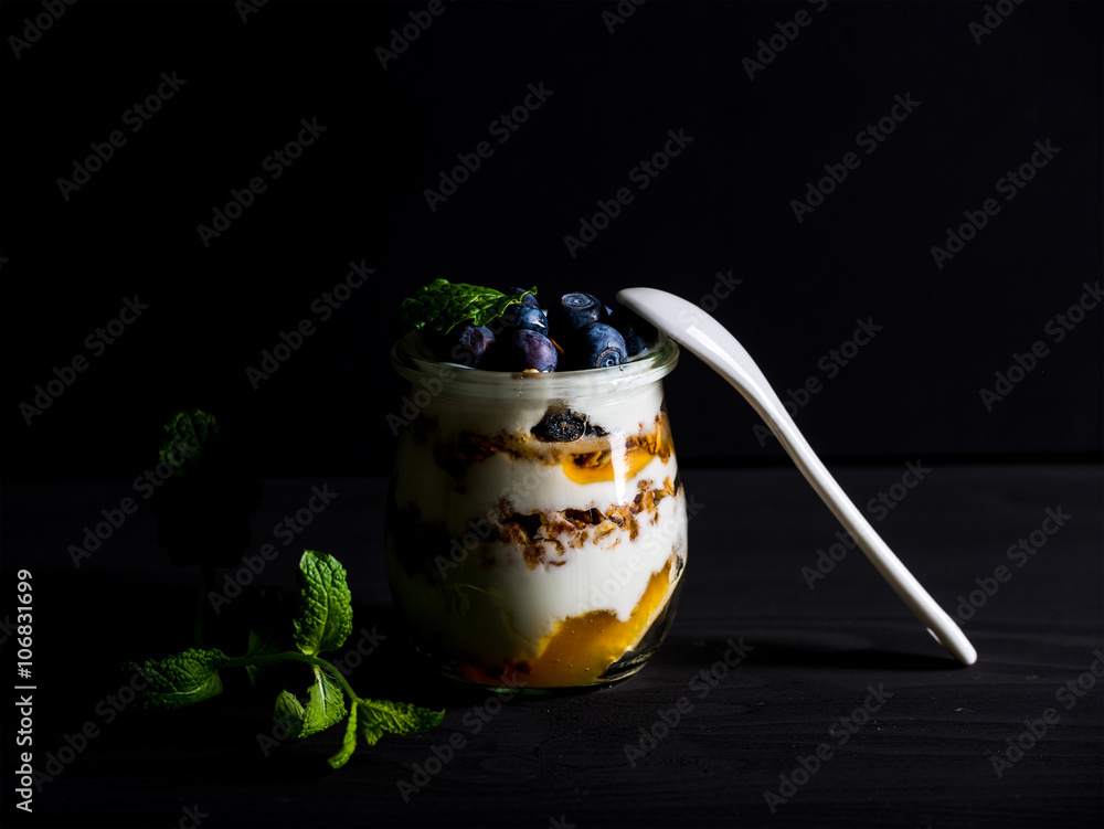 Yogurt oat granola with jam, blueberries and mint leavesin glass jar on black backdrop