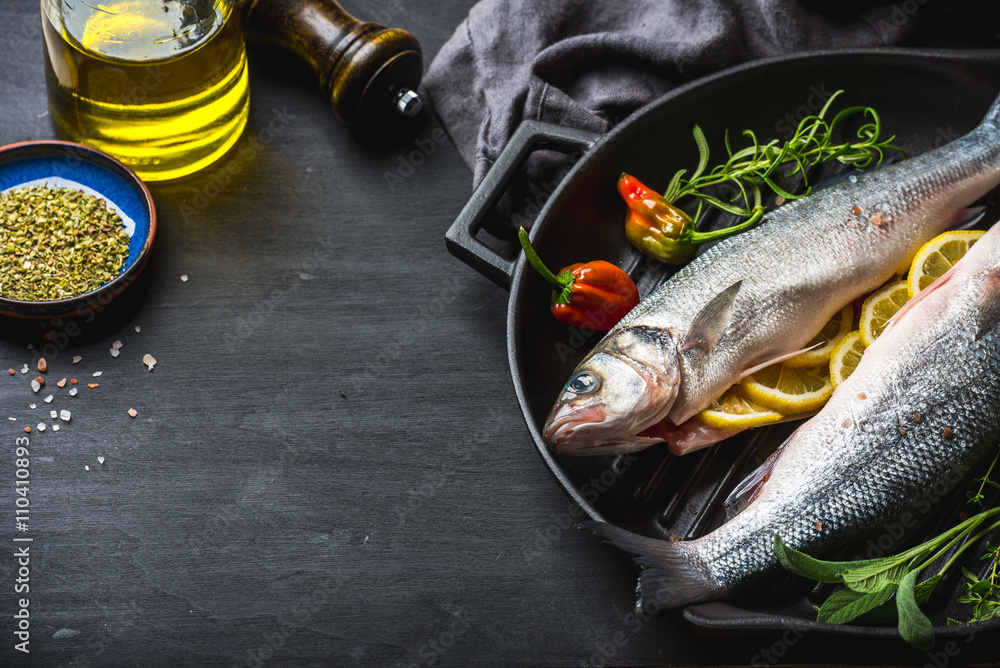 Raw uncooked seabass fish with herbs and spices in cast iron cooking pan on black wooden background