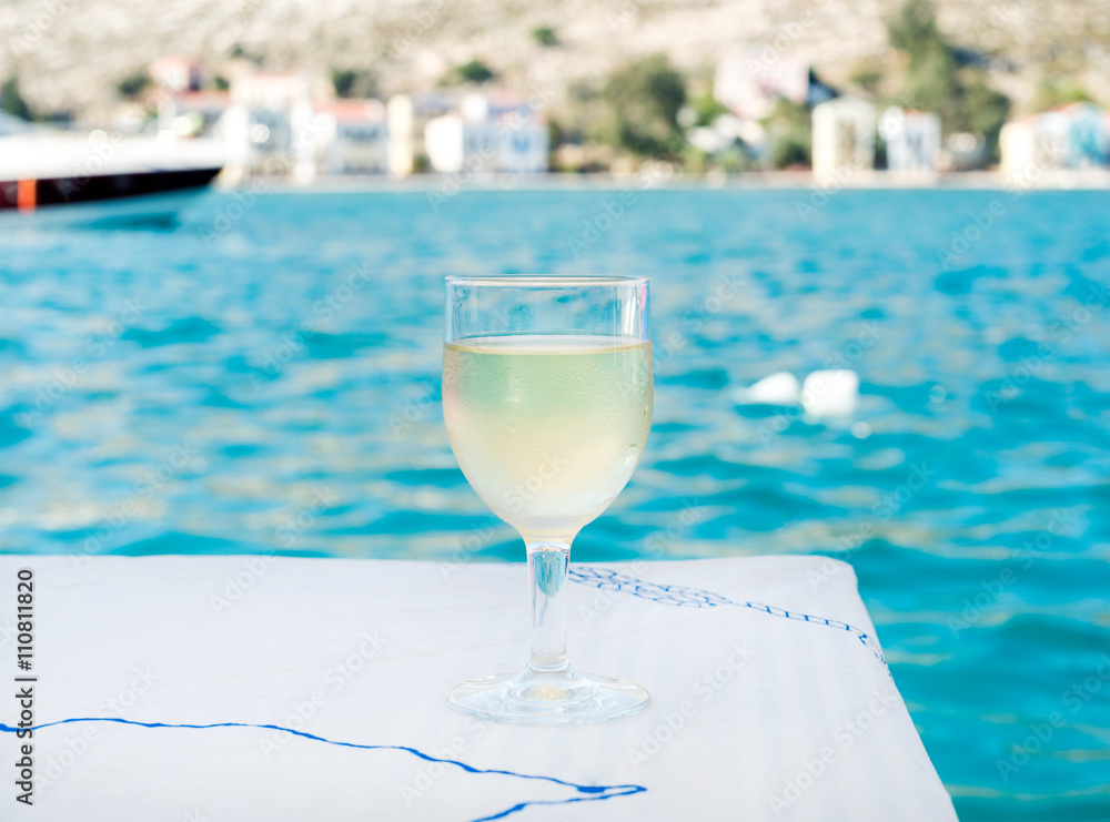 Glass of white wine on table in beach restaurant with sea view, blue water and yacht at background