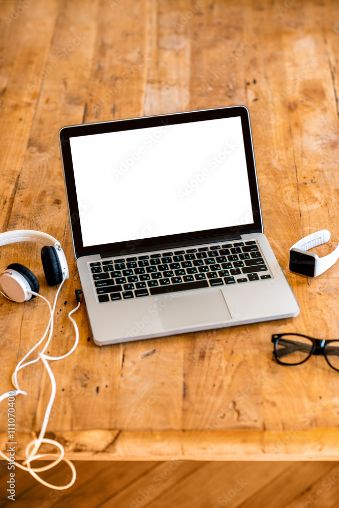 Laptop with white screen with earphones, smart watch, glasses on the wooden table