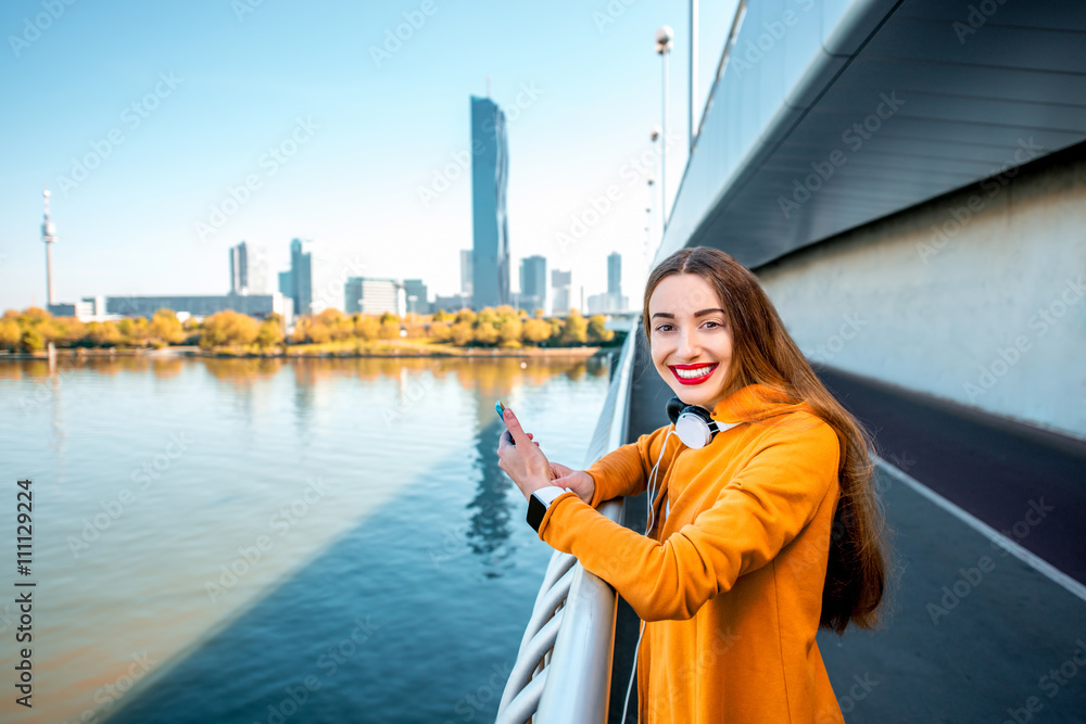 Young sport woman in yellow sweater using smart phone on the modern bridge with skyscrapers on the b