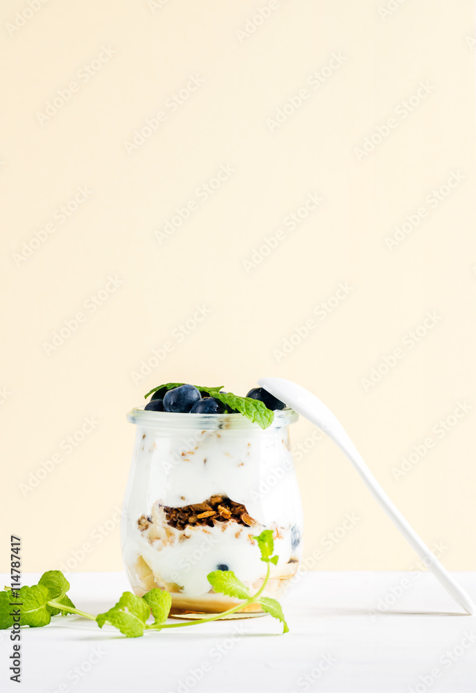 Yogurt oat granola with jam, blueberries and green leaves in glass jar on pastel yellow backdrop.