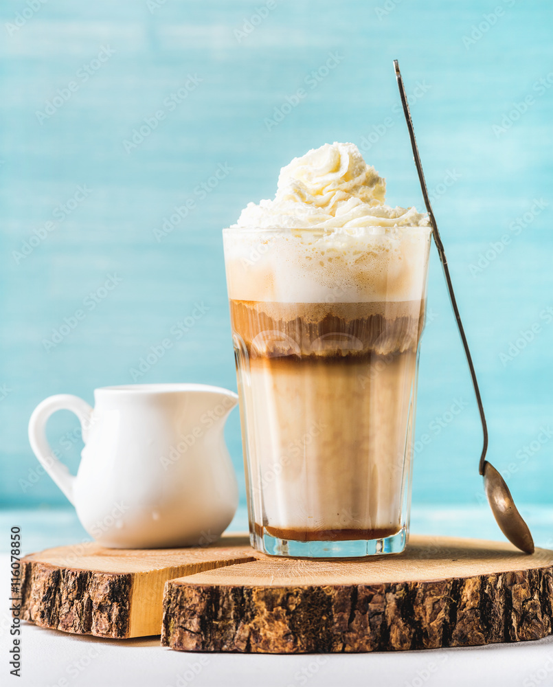 Latte macchiato with whipped cream, serving silver spoon and white pitcher on wooden round board ove