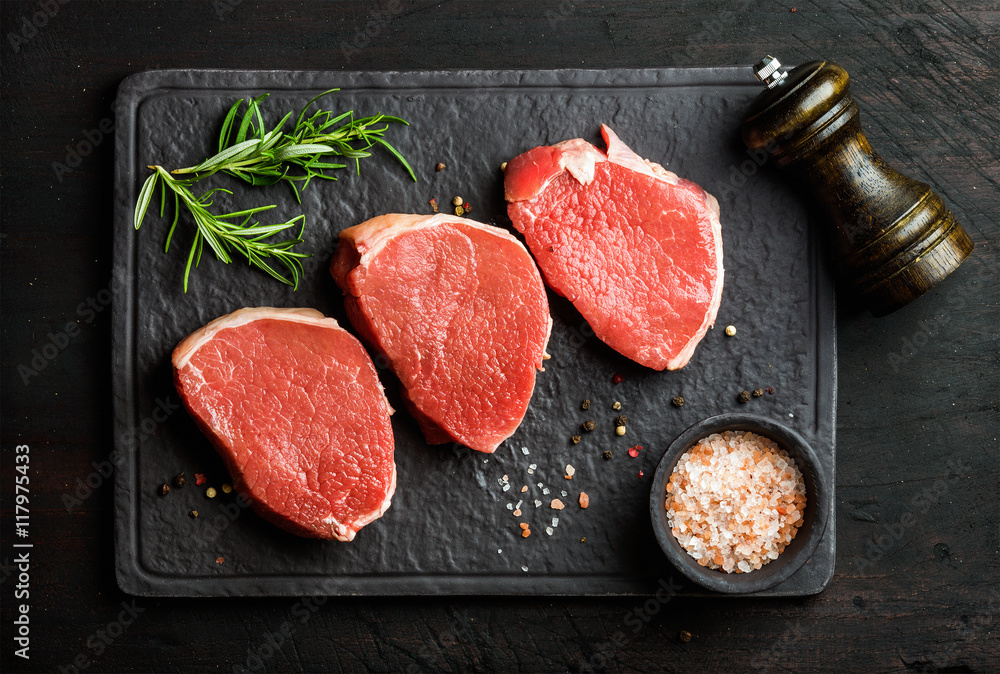 Raw beef Eye Round steaks with spices and rosemary on black slate stone board over dark wooden backg