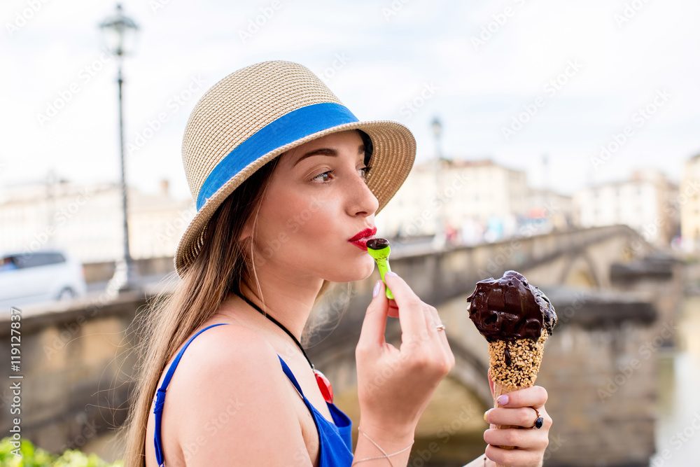 Young female tourist in blue dress eating chocolate ice cream in waffle cone sitting near the river 