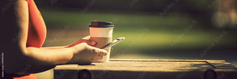 Woman holding smart phone and take away coffee in morning sunlight at the park