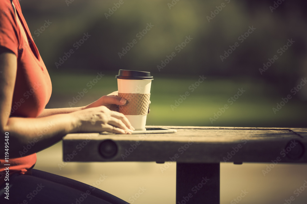 Woman holding smart phone and take away coffee in morning sunlight at the park