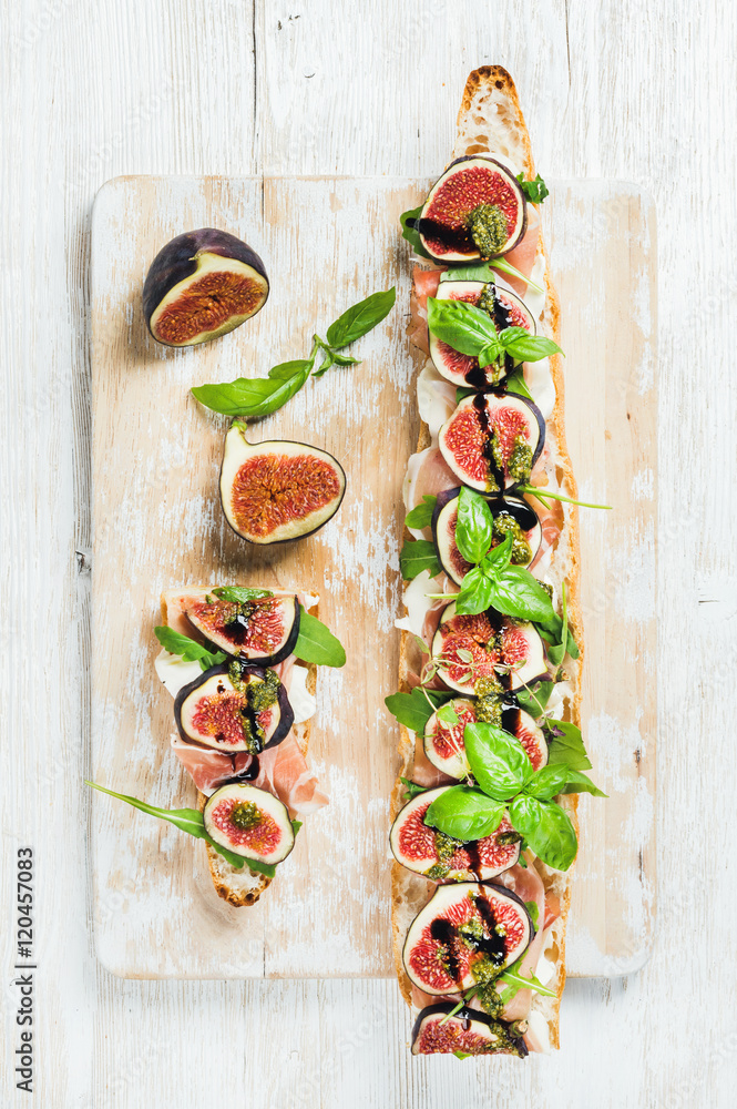 Long baguette sandwich with prosciutto meat, mozzarella cheese, arugula, figs and basil on shabby wh