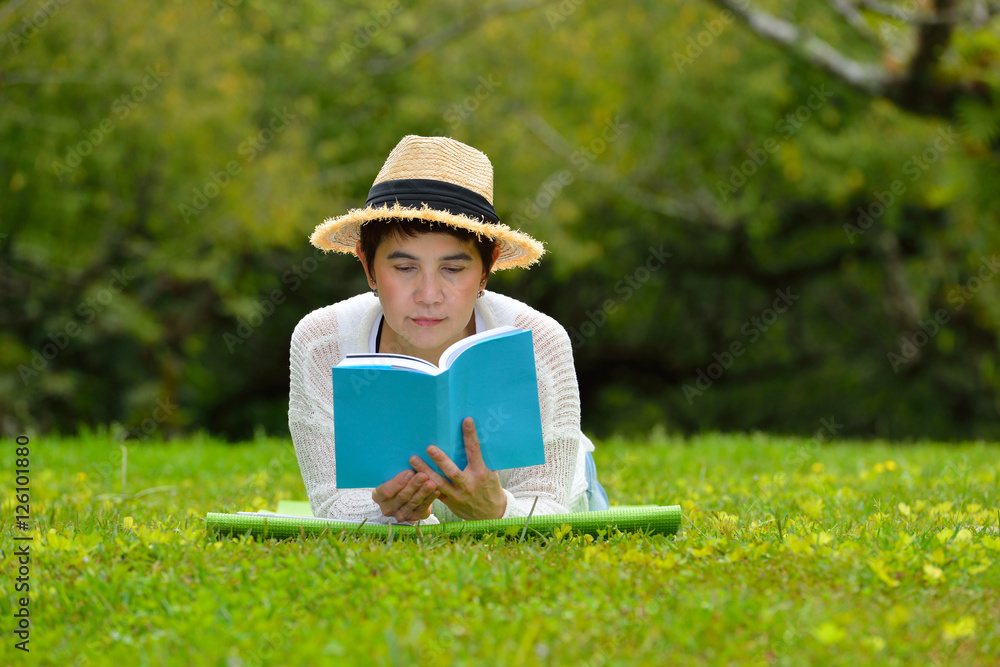 Happy middle aged woman lying on green grass reading a book in the park