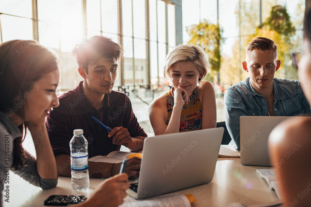 Group of students sitting at table in library and using laptop