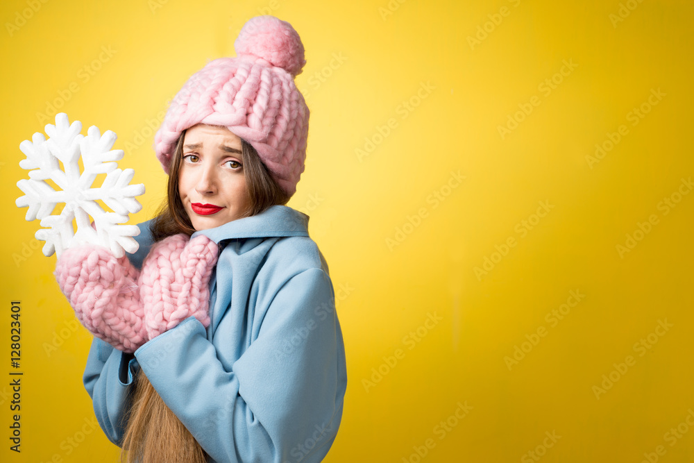 Confused and unhappy woman in colorful winter clothes holding a snowflake on the yellow background