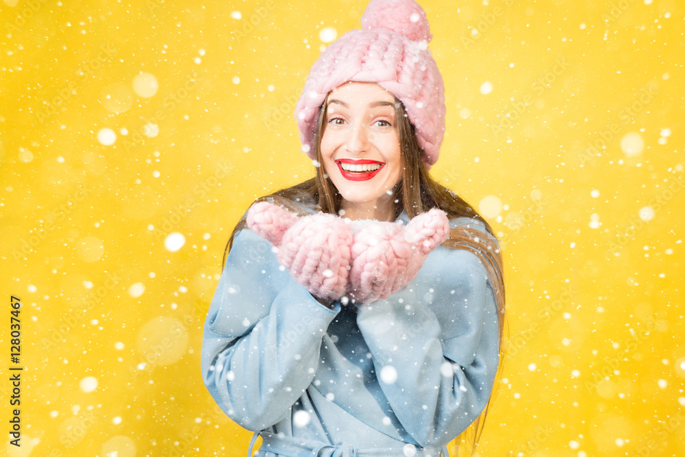 戴着针织帽子和手套的年轻女子在黄色的墙壁背景上抱着雪。五颜六色的幸福w