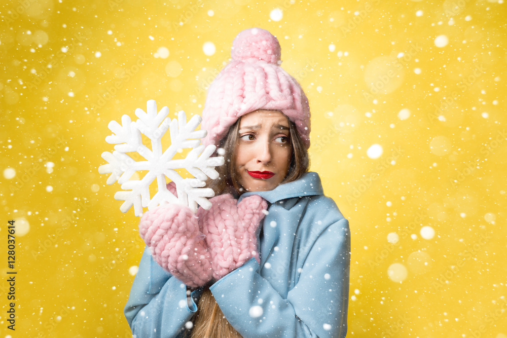 困惑而不快乐的女人穿着五颜六色的冬装，黄色背景上拿着一片雪花