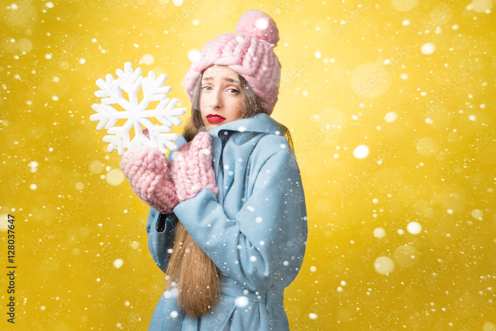 困惑而不快乐的女人穿着五颜六色的冬装，黄色背景上拿着一片雪花